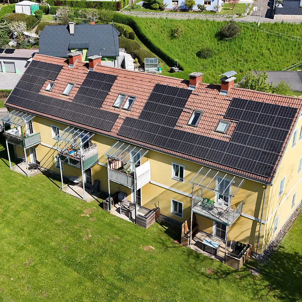 Luftaufnahme der Gemeinschaftsanlage Fohnsdorf-Kolonieweg. Die Anlagengröße beträgt 22,41 kWp und versorgt 8 Wohneinheiten. Die PV-Module sind auf dem Schrägdach eines Mehrparteienhauses installiert, das Teil einer Wohnungseigentümergemeinschaft (WEG) ist. Das Gebäude befindet sich in einer ruhigen, grünen Wohngegend mit umliegenden Einfamilienhäusern und weitläufigen Rasenflächen.